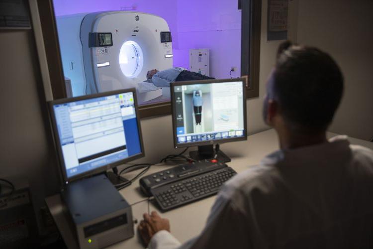 a medical professional looks through a window at a patient on a PET scanner while monitoring on screens